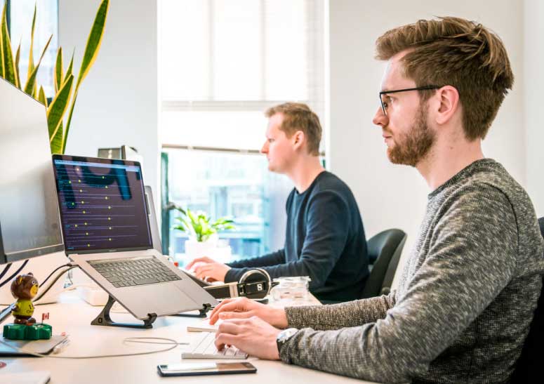 Two men working at a desk with laptops and monitors in a bright modern office.